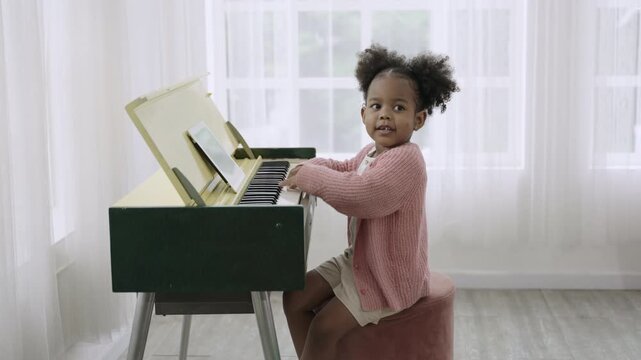 Happy cute African American childhood girl playing and learning to practice piano in classroom at nursery school. A little adorable black child showing skill in playing piano in living room at home. 