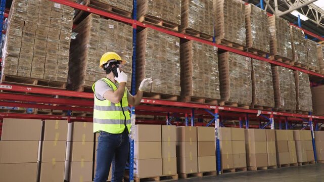 Young man warehouse worker in uniform wearing a yellow safety helmet and goggles, standing, using a smartphone, talking, discussing while checking inventory stock in a logistics distribution factory. 