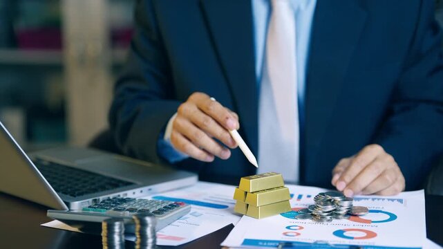 Businessman pointing pen between gold bars and coins to analyze and decide on investment allocation.