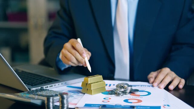 Businessman pointing pen between gold bars and coins to analyze and decide on investment allocation.