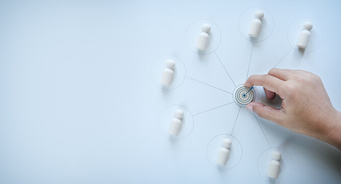 Hand placing central business target icon connected with wooden peg dolls representing employee team alignment, concept of corporate goal setting, business strategy communication, and team vision.