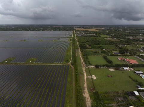 Aerial view of a large solar farm with rows of photovoltaic panels adjacent to green fields and rural properties under a cloudy sky in Miami, Florida, United States.