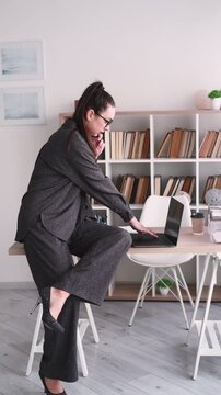 Vertical video. Business manager. Mobile communication. Busy woman talking on smartphone at office desk with laptop for paperwork indoor.