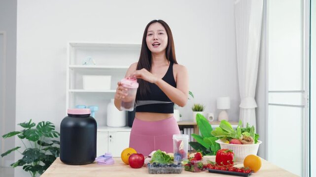 Asian digital creator sharing daily detox routine with organic fruits and blender. Active young woman live streaming fitness journey and nutritional advice from home kitchen.