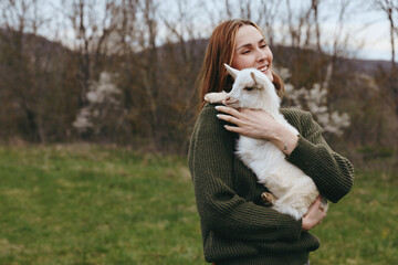 Naklejka premium A joyful woman in a knitted sweater hugs a white goat kid in a grassy countryside, capturing a simple lifestyle moment outdoors, with soft light and fresh air. The scene conveys affection and bond