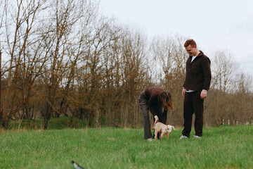 Fototapeta premium Casual lifestyle scene with two adults in casual clothes in a grassy field, bending to greet a small light dog, during an autumn afternoon in a tranquil park setting, with soft natural light leaves