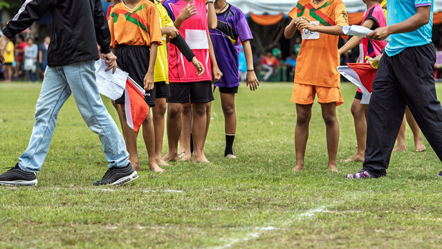 Track referee holding signal flags at school sports day. Sports referee holding red and white signaling flags and competition documents, standing on grass field track surrounded by young schoolgirls.