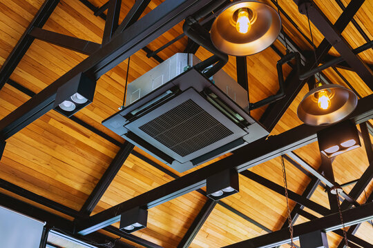 Modern cassette type air conditioner installed on loft ceiling with steel beams and wooden roof. Ceiling-mounted cassette air conditioning unit integrated into modern interior design. HVAC systems.