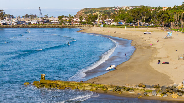Aerial view of Dana Point beach with surfers carrying boards, calm blue waves, sandy shoreline, palm trees, and harbor infrastructure in Dana Point, California, United States.