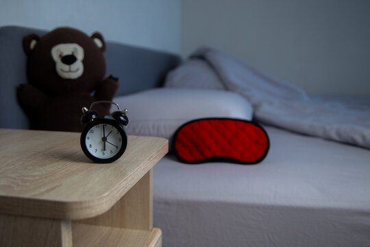 There is a black mini alarm clock on a wooden bedside table, and in the background on a gray bed is a red sleep mask and a brown teddy bear. The concept of early awakening, healthy sleep, sleep regime