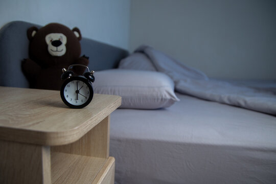 There is a wooden bedside table with a black alarm clock in the foreground, and a brown teddy bear and gray pastels in the background. The concept of early awakening, healthy sleep, sleep patterns