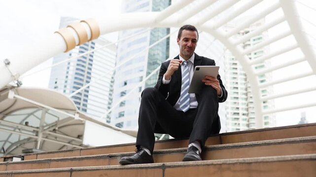 Confident businessman in a suit sits on city steps with a tablet, celebrating success against modern skyscrapers and a white architectural skywalk.