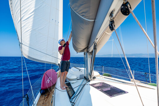 Man trimming mainsail on a sailing yacht during offshore voyage. Experienced sailor adjusting sail shape for efficient wind propulsion, safe navigation, boat control and blue water cruising lifestyle.