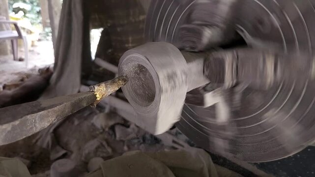 Slow-motion horizontal footage of an Indian artisan shaping a granite stone utensil on a rotating lathe machine. The spinning stone is carefully worked with cutting tools to create a rounded form