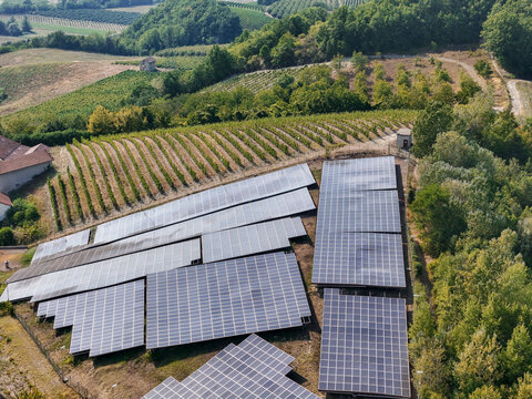 Aerial view of a solar power plant installed on a hilltop, replacing a section of a vineyard surrounded by lush green hills and trees in Monastero Bormida, Piedmont, Italy.