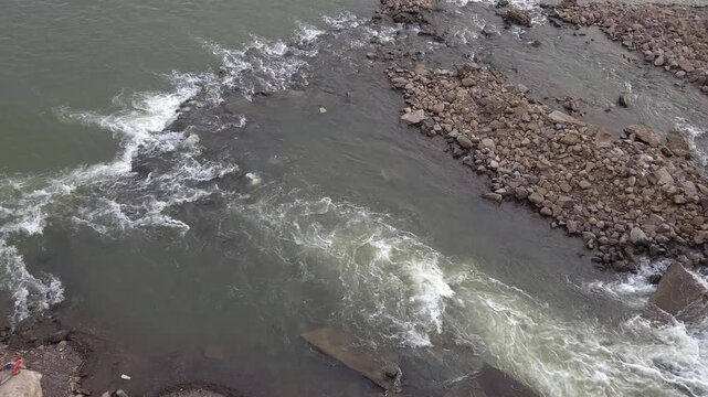 A wide turbulent riverbed channel from open stone shores to the foundational base of a concrete dam structure, illustrating environmental engineering and heavy water runoff management