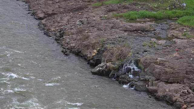 Rocky Shoreline and Running River Water with Small Stream Flowing into Main Current in Outdoor Nature Setting.