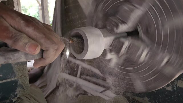 Slow-motion horizontal footage of an Indian artisan shaping a granite stone utensil on a rotating lathe machine. The spinning stone is carefully worked with cutting tools to create a rounded form