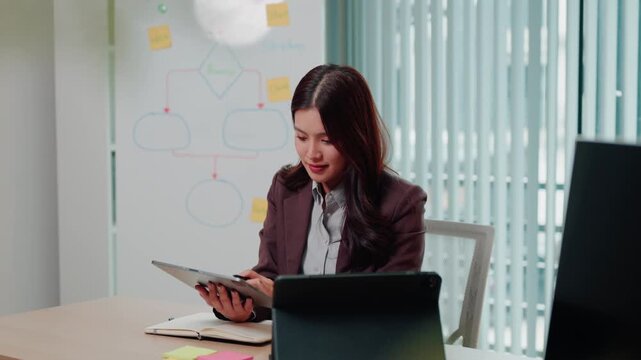 Asian Businesswoman Using Digital Tablet at Office Desk with Whiteboard Flowchart in Background