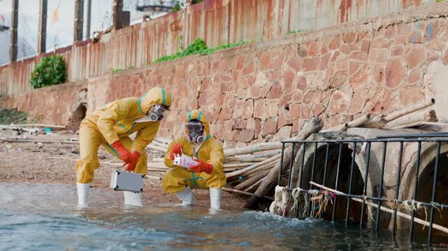 Two technicians in hazmat suits collect water samples at a drainage outfall, testing pollution and contamination along a flooded urban shoreline.