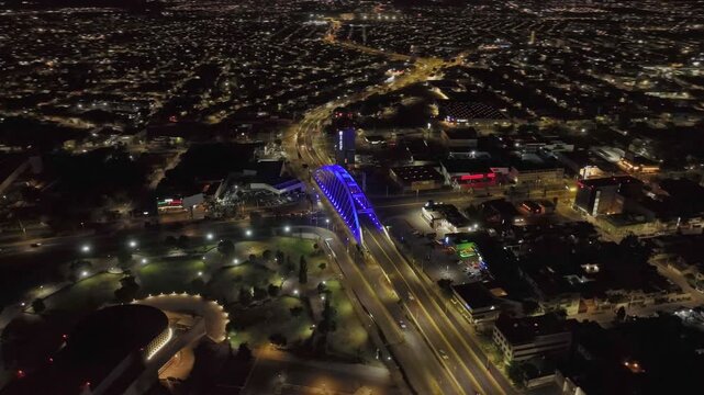 Wide aerial orbit over Aguascalientes at night, following Avenida Aguascalientes Sur through the city with the blue illuminated Bicentenario Bridge as its visual centerpiece.