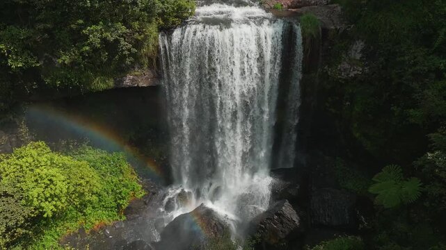 Aerial view of Zillie Falls cascading into a lush rainforest pool with a vibrant rainbow visible in the mist in Millaa Millaa, Queensland, Australia.
