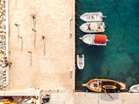 Aerial view of boats moored at the stone pier with people walking on the paved dock in the clear turquoise water of the harbor in Brela, Split-Dalmatia County, Croatia.