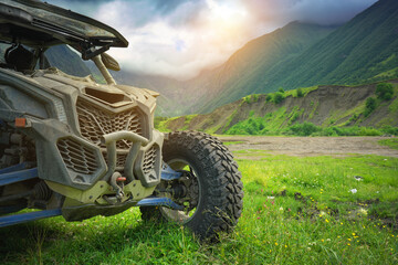 Сlose up of a yellow dirty offroad UTV buggy car parked on a dirt trail looking towards a majestic green mountain valley under a cloudy sky. Extreme motorsport travel adventure concept design © GIS