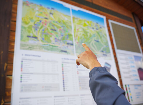 Person&rsquo;s hand pointing at a detailed ski resort map mounted on a wooden wall, featuring color‑coded trails, elevation zones