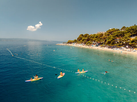 Aerial view of people paddleboarding on the clear turquoise waters of Punta Rata beach, surrounded by lush pine trees and the Adriatic Sea in Brela, Split-Dalmatia County, Croatia.