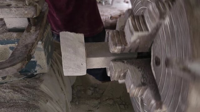 Slow-motion horizontal footage of an Indian artisan shaping a granite stone utensil on a rotating lathe machine. The spinning stone is carefully worked with cutting tools to create a rounded form