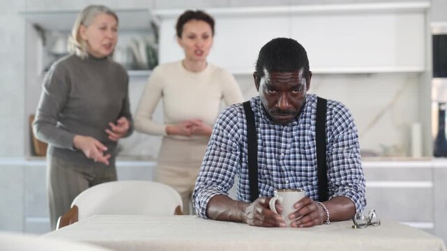 Sad middle-aged man sitting at the kitchen table with his back to old and middle-aged women quarreling to him 