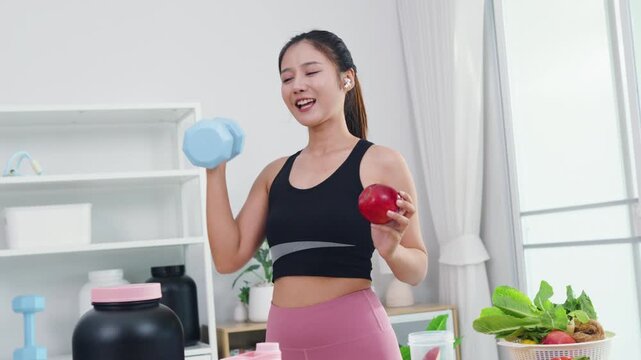 Asian fitness woman holding dumbbell and protein shake for home workout routine. Sporty female influencer showing sports nutrition supplements and healthy organic lifestyle in modern kitchen.