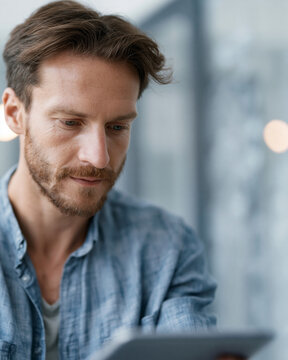 Man with beard focuses on tablet screen. Casual denim shirt, thoughtful expression. Soft lighting creates calm, modern atmosphere. Ideal for business, education, or tech-related topics