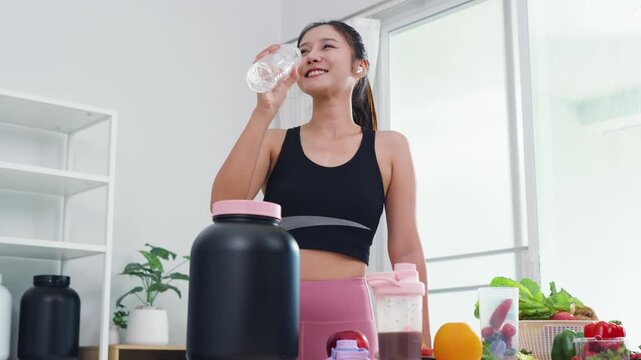 Asian fitness woman holding dumbbell and protein shake for home workout routine. Sporty female influencer showing sports nutrition supplements and healthy organic lifestyle in modern kitchen.