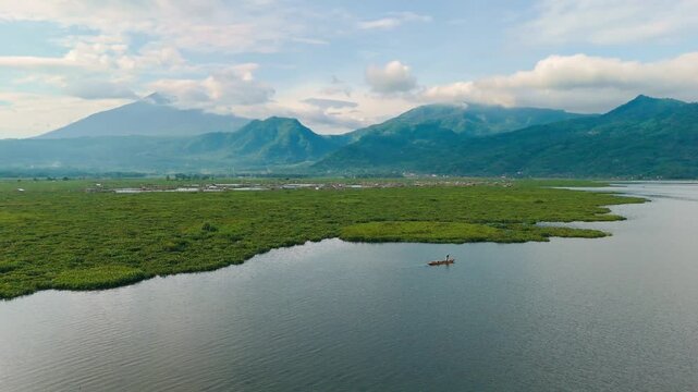Aerial view of a lone fisherman in a small boat on Lake Rawa Pening surrounded by water hyacinth and distant mountains in Waduk Rawapening, Central Java, Indonesia.