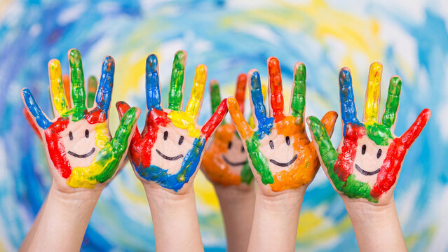 Children's hands covered in colorful paint with smiley face drawings are held up against a painted artistic canvas background.