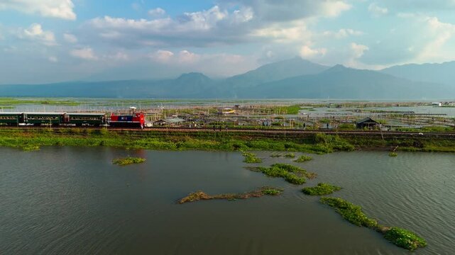 Aerial view of a train crossing the railway tracks on Lake Rawa Pening with lush mountains in the background under a clear sky in Waduk Rawapening, Central Java, Indonesia.