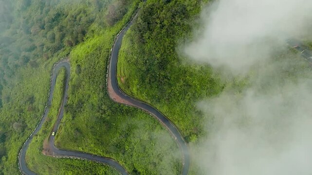 Aerial view of a winding road with vehicles driving towards the peak of Mount Telomoyo surrounded by lush green forests and clouds in Ngablak Regency, Central Java, Indonesia.