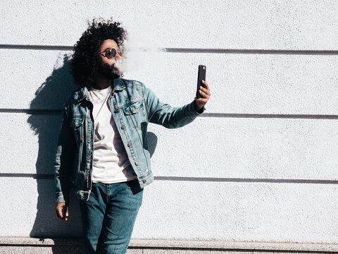 Handsome smiling hipster model.Unshaven Arabian man smoking electronic cigarette. Fashion male with long curly hairstyle posing in the street. Makes smoke. Steam from vape. Takes selfie
