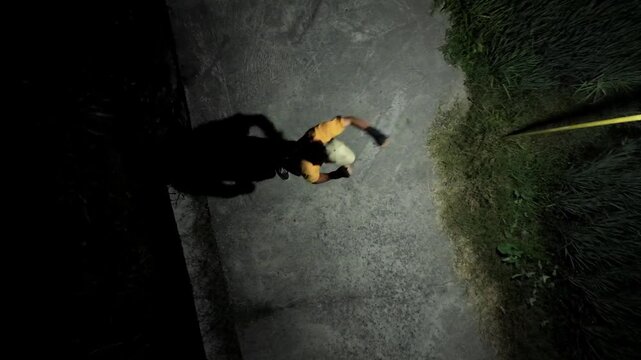 Slow motion drone ascends and rotates revealing a top-down aerial view of a man in a cap shadow boxing at night. He trains martial arts on an outdoor path under dramatic artificial lighting.