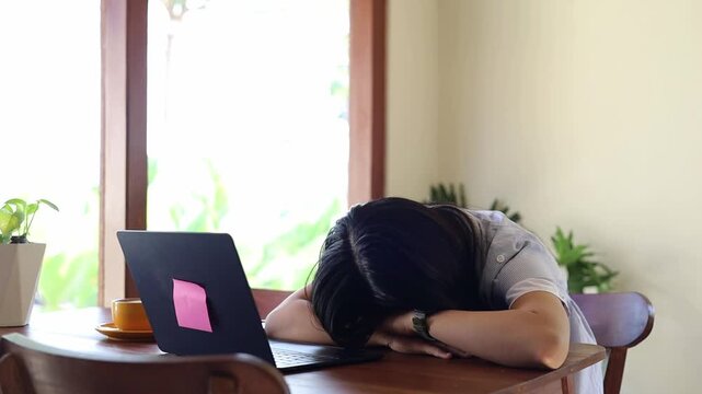 Young Asian woman sleeping on desk due to overworking and exhausting while working on laptop in cafe