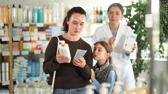 Interested woman reading label on baby cream package for preteen daughter, focusing on natural ingredients, while friendly female pharmacist providing guidance on safe child skincare at drugstore 