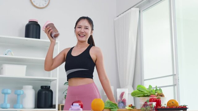 Smiling Asian girl showing wellness lifestyle and healthy fitness routine at home. Happy energetic woman preparing organic diet with fresh vegetables and protein shaker for daily wellbeing.