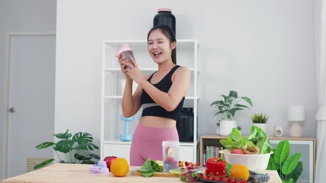 Smiling Asian girl showing wellness lifestyle and healthy fitness routine at home. Happy energetic woman preparing organic diet with fresh vegetables and protein shaker for daily wellbeing.