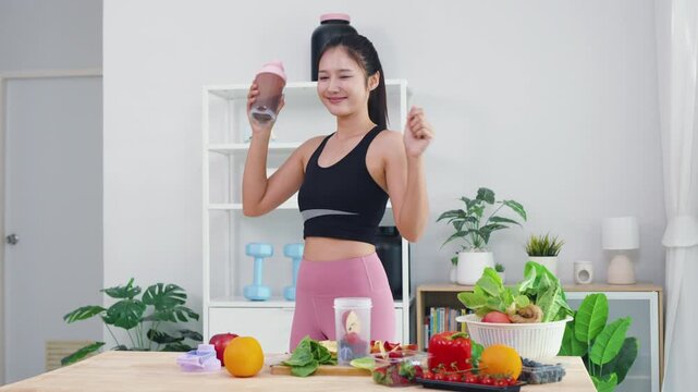 Smiling Asian girl showing wellness lifestyle and healthy fitness routine at home. Happy energetic woman preparing organic diet with fresh vegetables and protein shaker for daily wellbeing.