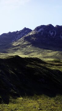 Rolling green hills stretch across the landscape, embraced by towering snow capped mountains under a clear blue sky. Morning sunlight casts gentle shadows, creating a serene atmosphere.