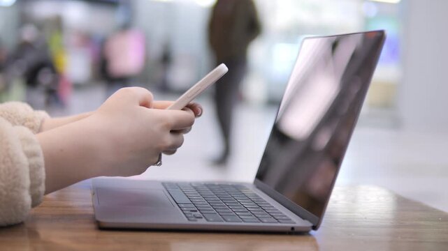 Hands hold smartphone near open laptop on wooden table. Background shows blurred indoor public space. Person wears light-colored sleeve. Focus remains on hands and devices. No animals or plants shown