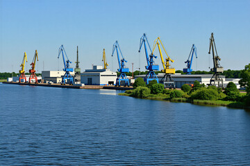 Fototapeta premium Large port cranes stand by water. Industrial dockyard has facilities and warehouses. Blue sky over maritime transport hub. Ships load and unload cargo.