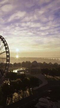 Morning light blankets the amusement park, revealing a giant wheel amidst lush trees. Soft shadows emerge as day breaks, creating a serene and inviting scene.